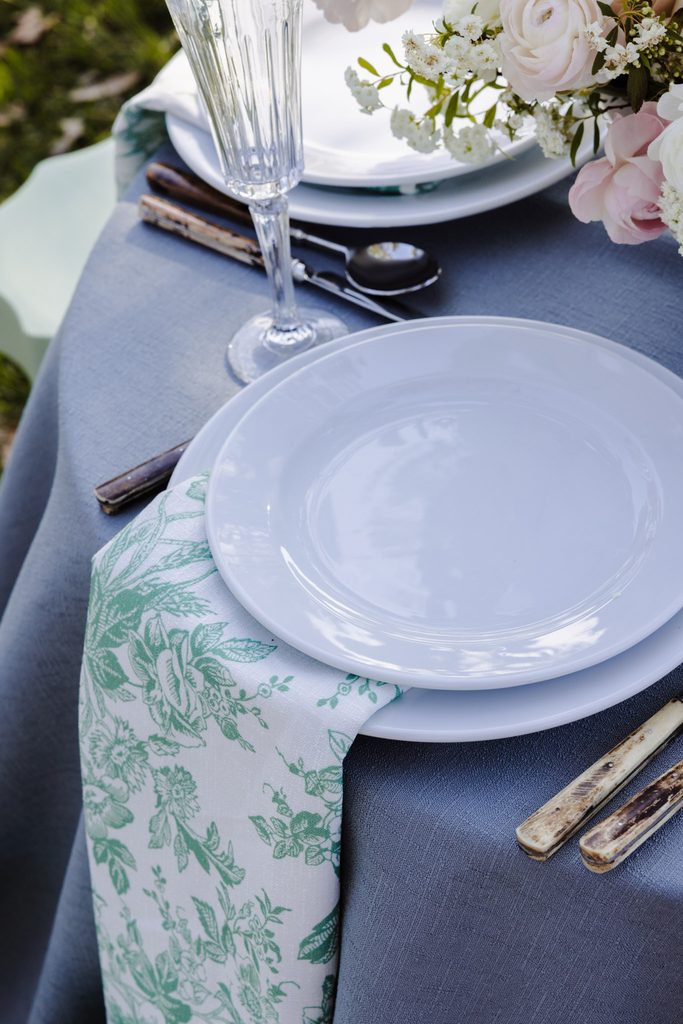 A place setting with white plates, a champagne flute, silverware, and a green and white floral napkin arranged on a blue Emma Blomfield rug.