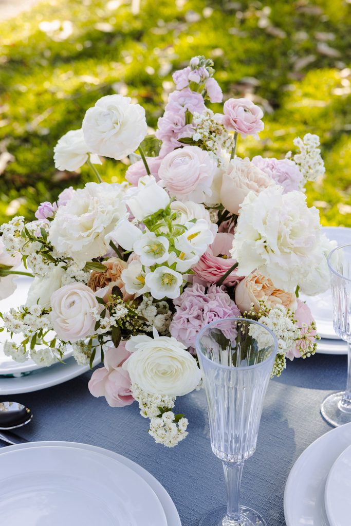 A floral centerpiece of white and pale pink blooms sits on a table with white plates and glasses atop an Emma Blomfield rug.
