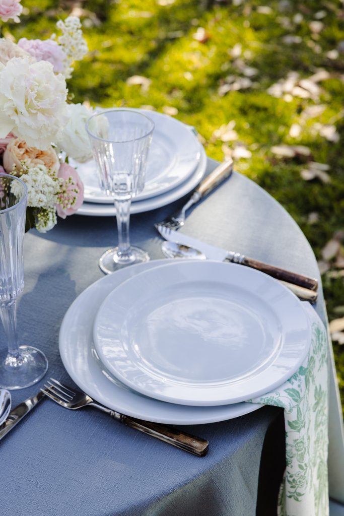 A place setting with white plates, glassware, and flatware on a gray Emma Blomfield rug next to a floral centerpiece.