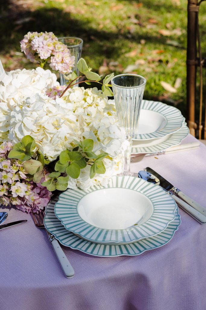 A table setting on an Emma Blomfield rug features a lavender cloth, white and green striped plates, a tall glass, and a large bouquet of white and pink hydrangeas.