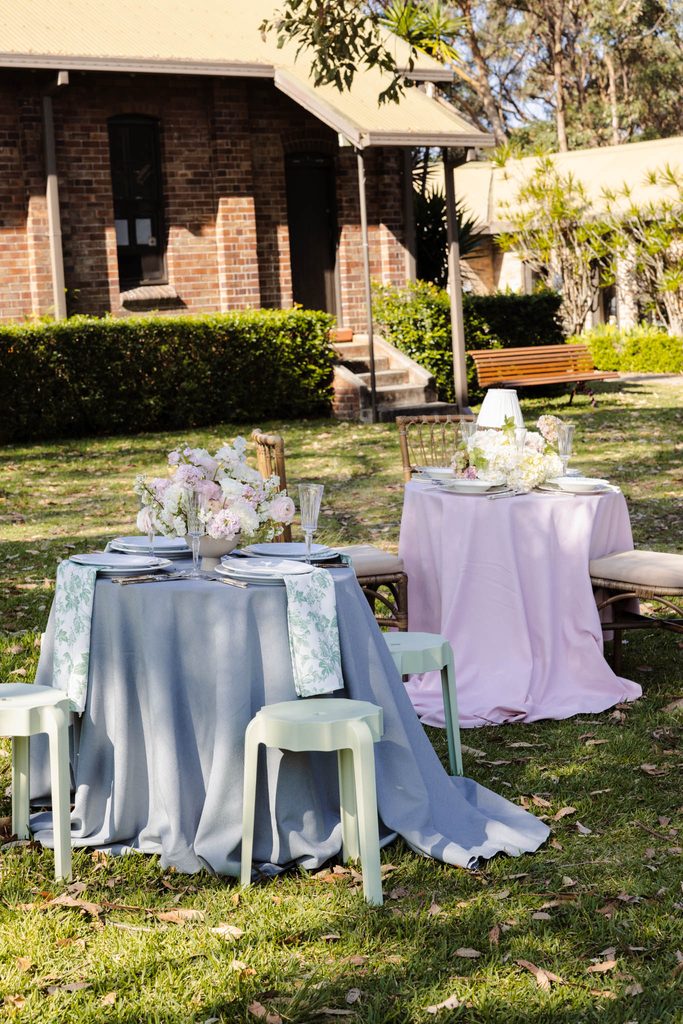 Two round tables with floral centerpieces set for outdoor dining on a grassy lawn styled with an Emma Blomfield rug collection.