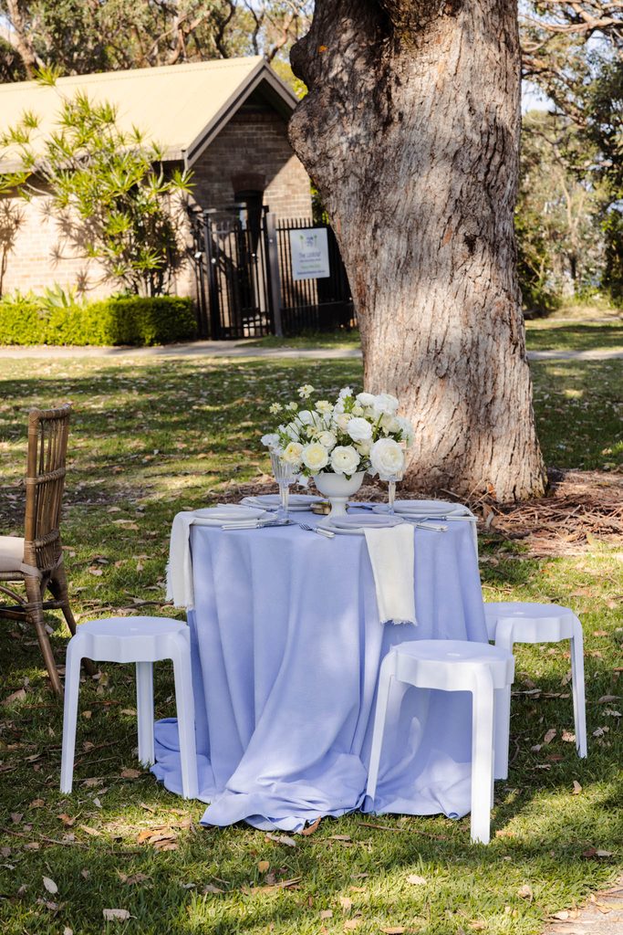 A small round table covered in a light blue cloth, set for four with a white floral centerpiece, stands on an Emma Blomfield rug on a grassy lawn.