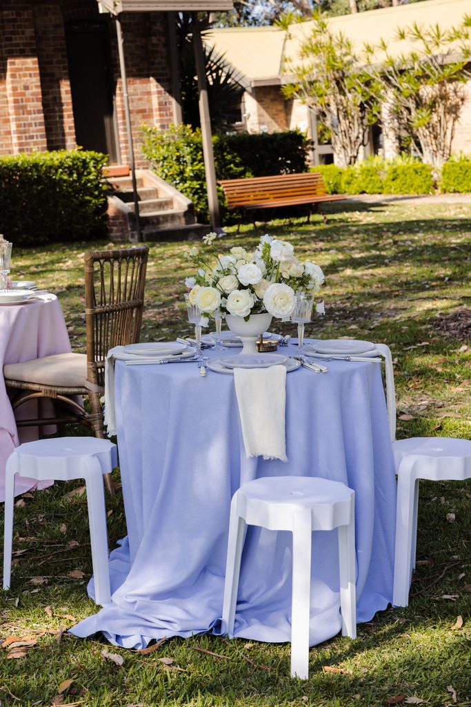 A small round table with a blue tablecloth, white floral centerpiece, and white stools set up on an Emma Blomfield rug on the lawn.
