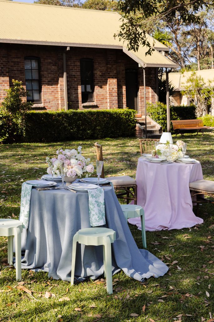 Emma Blomfield sits at one of two round tables featuring floral centerpieces and place settings from the Emma Blomfield rug collection on a grassy lawn in front of a brick building.