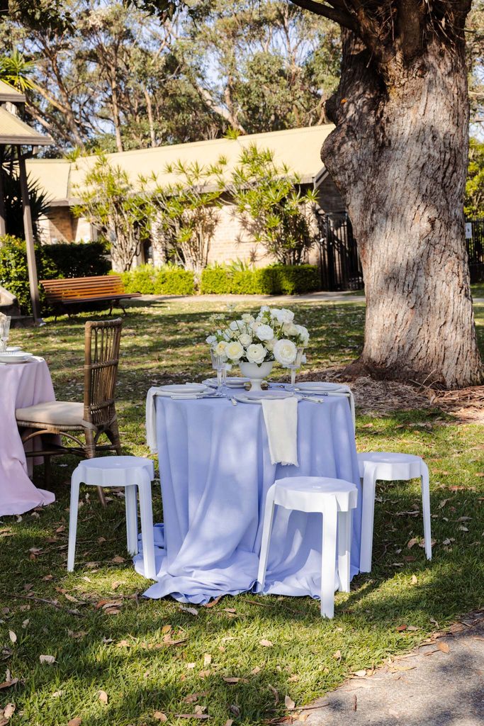 A round table with a lavender cloth and white floral centerpiece sits on an Emma Blomfield rug surrounded by three white stools on a grassy lawn.