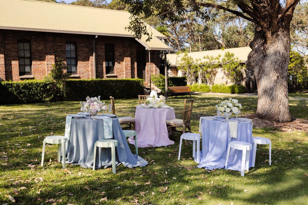 Three small round tables with floral centerpieces set on an Emma Blomfield rug collection on a lawn in front of a brick building.