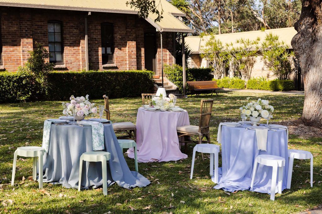 Emma Blomfield stands near three round tables with pastel tablecloths and floral centerpieces set up on a lawn in front of a brick building featuring her rug collection.