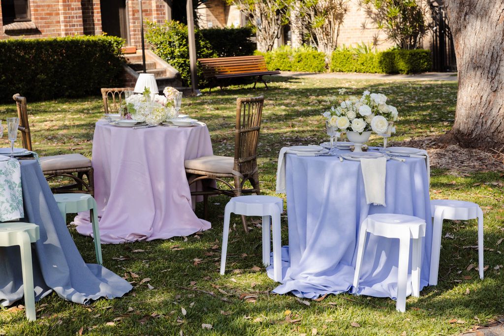 Small round tables with pastel tablecloths and floral centerpieces are set up on an Emma Blomfield rug on a grassy lawn.