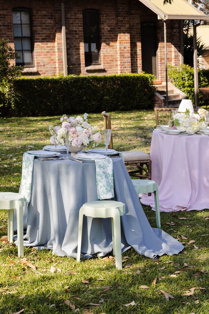 Two round tables with Emma Blomfield rugs and floral centerpieces are set up on a lawn for an outdoor event in front of a brick building.