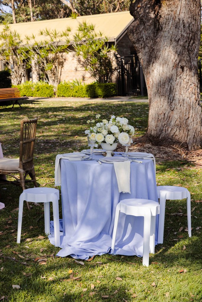 A round table covered in a light blue Emma Blomfield rug cloth, set for two with a white floral centerpiece, surrounded by three stools in a park setting.