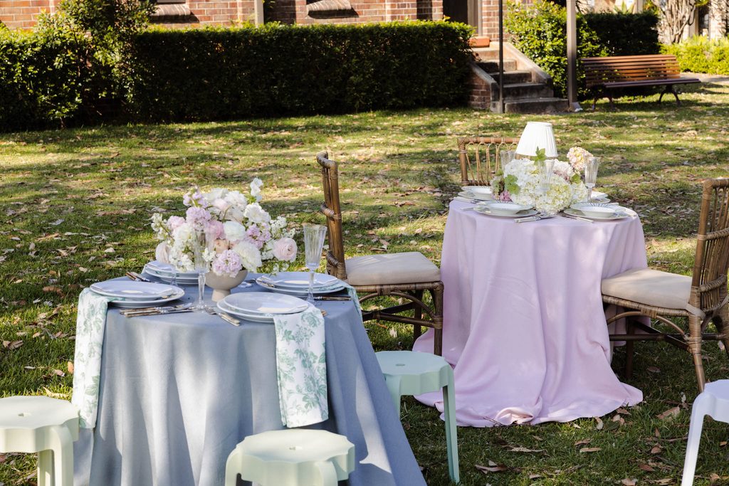 Emma Blomfield stands by two round tables with floral centerpieces on a lawn, styled with an Emma Blomfield rug collection for a garden event.