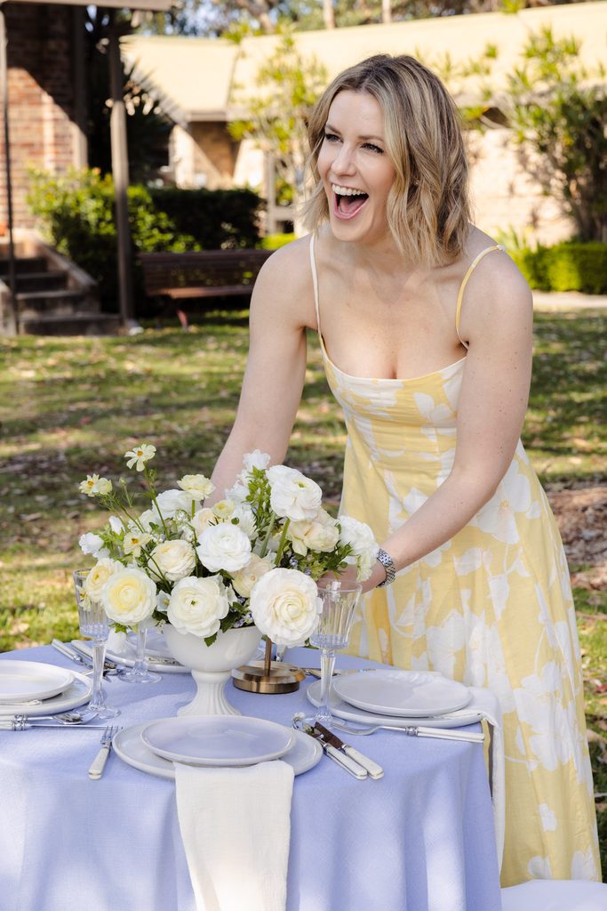 Emma Blomfield in a yellow floral dress arranges a floral centerpiece on an outdoor dining table styled with an Emma Blomfield rug.