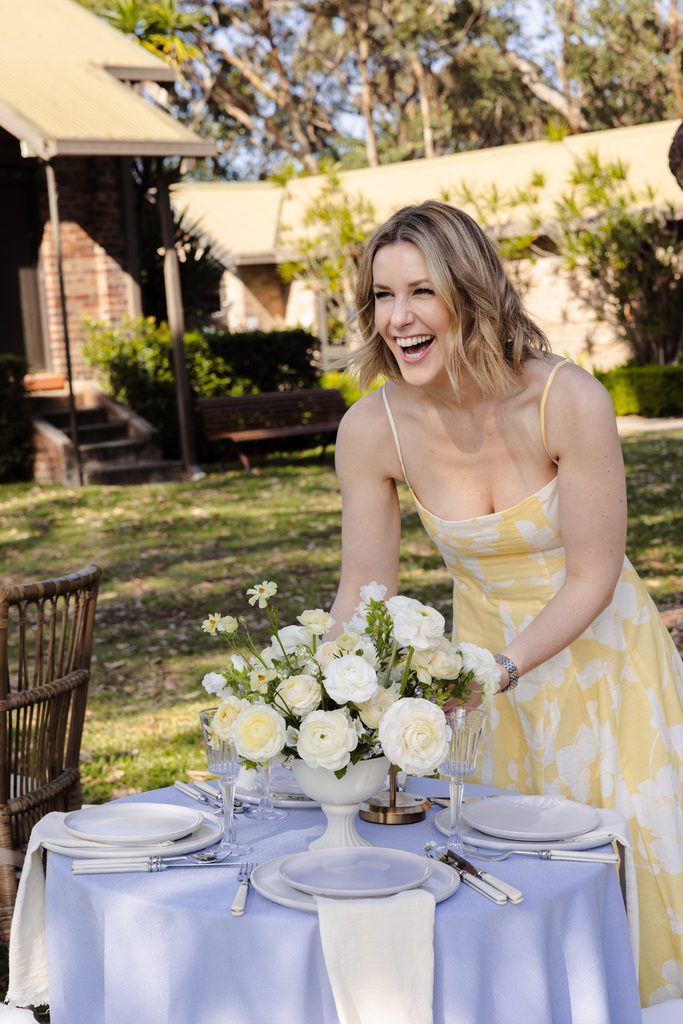 Emma Blomfield, in a yellow floral dress, sets a table with a white flower centerpiece in a garden on an Emma Blomfield rug.