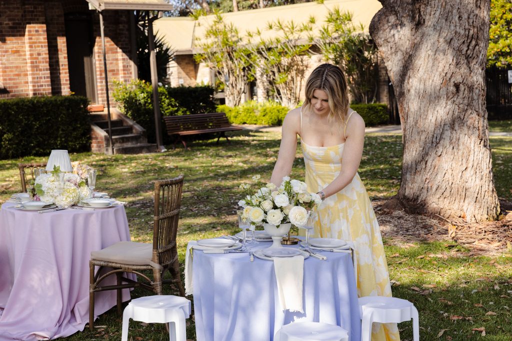 Emma Blomfield places a floral centerpiece on a table set for a garden event, grounding the space with an Emma Blomfield rug.